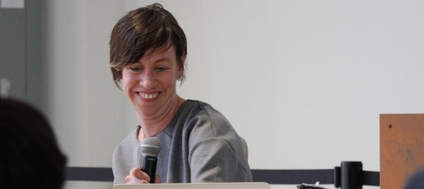 Eliza Chandler, a white disabled woman with short brown hair, sits smiling, looking down at a laptop on a table, holding a microphone. She appears to be in a classroom with people out of focus in the foreground.
